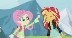 Size: 1363x727 | Tagged: safe, screencap, fluttershy, sunset shimmer, equestria girls, friendship through the ages, g4, alternate clothes, alternate hairstyle, blowing, braid, clothes, dandelion, female, flower, folk fluttershy, jacket, mountain, vest