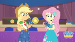 Size: 1057x594 | Tagged: safe, screencap, applejack, fluttershy, best in show: the victory lap, equestria girls, g4, my little pony equestria girls: better together, female, looking at you, microphone, smiling