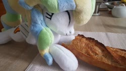 Size: 1207x679 | Tagged: safe, artist:ketika, vapor trail, pegasus, pony, g4, baguette, bread, cute, eating, eyes closed, food, happy, irl, kitchen, lying down, photo, plushie, solo, table, vaporbetes
