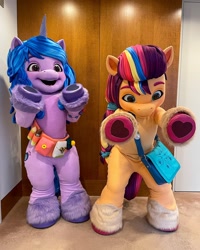 Size: 1080x1350 | Tagged: safe, izzy moonbow, sunny starscout, earth pony, human, pony, unicorn, g5, official, 2022, bag, belt, fursuit, irl, irl human, mascot, mascot costume, outstretched arms, photo, ponysuit, raised hoof, smiling, wide eyes, wide hips