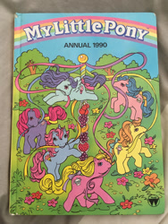 Size: 600x800 | Tagged: safe, official comic, banana surprise, coco berry, crunch berry, peppermint crunch, sherbet (g1), swirly whirly, earth pony, pegasus, pony, unicorn, g1, my little pony annual 1990, book cover, bow, cover, cover art, female, flower, horn, irl, mare, maypole, mouth hold, my little pony logo, photo, smiling, spread wings, tail, tail bow, tree, wings