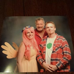 Size: 960x960 | Tagged: safe, big macintosh, fluttershy, human, g4, 2015, clothes, cosplay, costume, cutie mark on clothes, dress, female, floral head wreath, flower, holding hands, irl, irl human, john de lancie, looking at you, male, photo, plaid shirt, popcon, ship:fluttermac, shipping, shirt, smiling, smiling at you, straight, t-shirt, trio, wig, wings