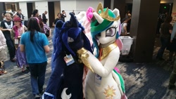 Size: 960x540 | Tagged: safe, princess celestia, princess luna, alicorn, human, g4, convention, crowd, duo focus, female, fursuit, indoors, irl, irl human, jewelry, looking at you, photo, ponysuit, regalia, royal sisters, siblings, sisters